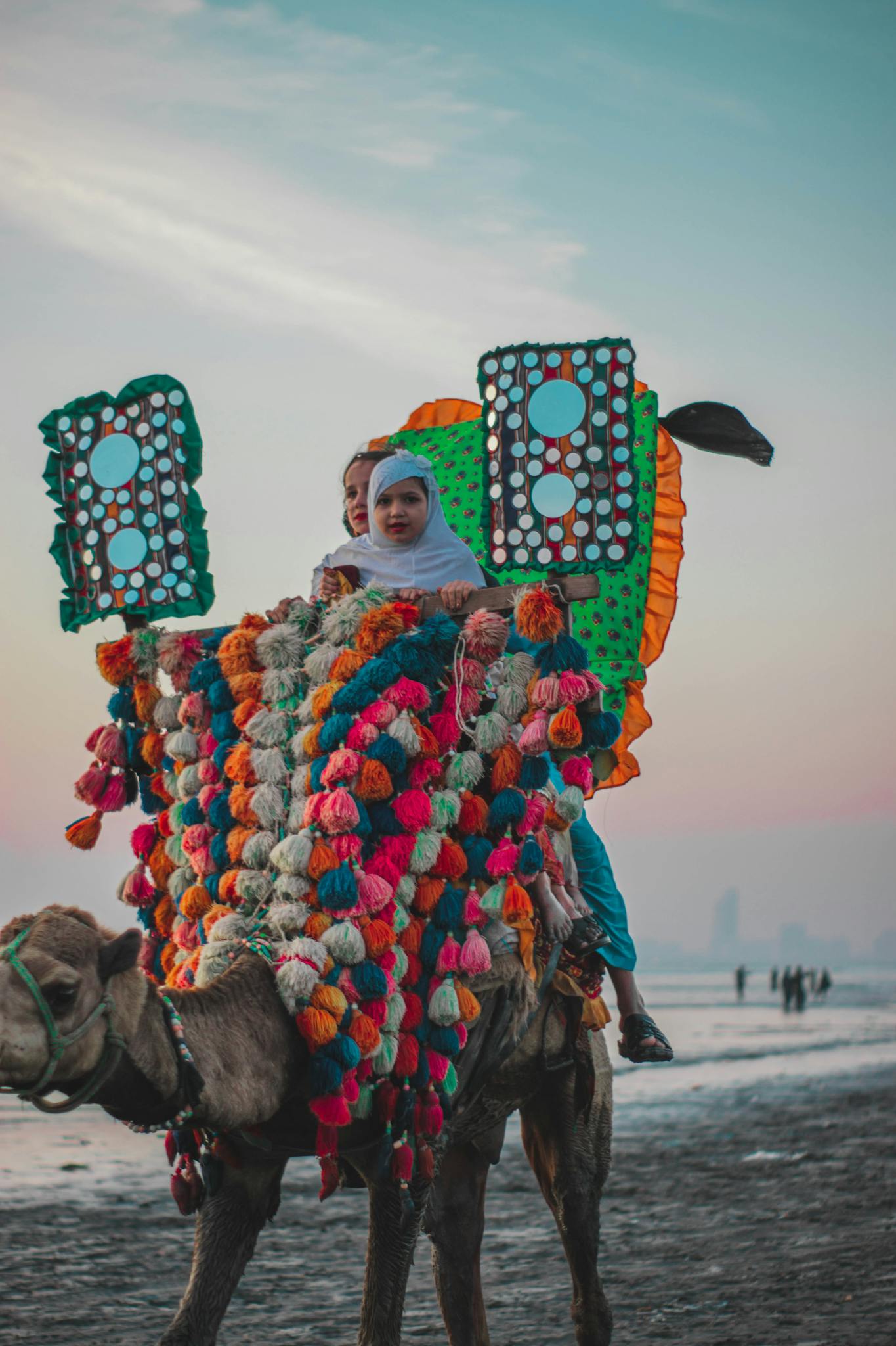 Two people riding a decorated camel on a beach during sunset with vibrant colors.